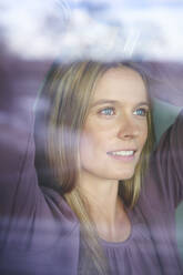 Porträt einer lächelnden blonden jungen Frau hinter einer Fensterscheibe - PNEF02432