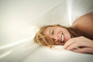 Porträt einer glücklichen blonden jungen Frau im Bett liegend unter einer Decke - PNEF02445
