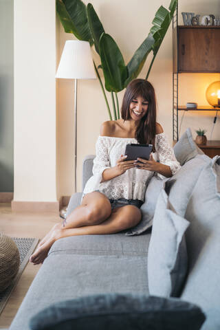 Glückliche junge Frau, die zu Hause auf der Couch sitzt und ein Tablet benutzt, lizenzfreies Stockfoto