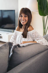 Portrait of smiling young woman sitting on the couch at home - MPPF00522