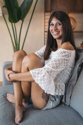 Porträt einer lächelnden jungen Frau, die zu Hause auf der Couch sitzt - MPPF00524