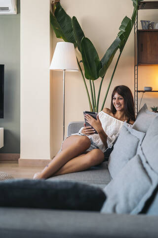 Lächelnde junge Frau sitzt auf der Couch zu Hause mit Tablet, lizenzfreies Stockfoto