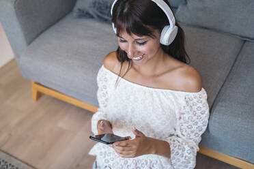 Happy young woman listening to music with headphones and sitting on the floor in the living room at home - MPPF00527