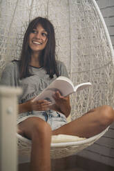 Smiling young woman sitting in hanging chair reading a book - MPPF00533