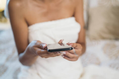 Clos-up of young woman wrapped in a towel sitting on bed holding cell phone stock photo