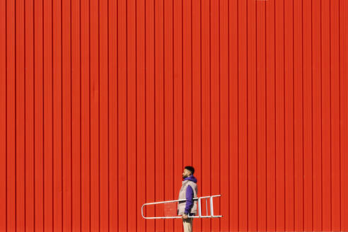 Young man carrying a ladder in front of a red wall - ERRF02821