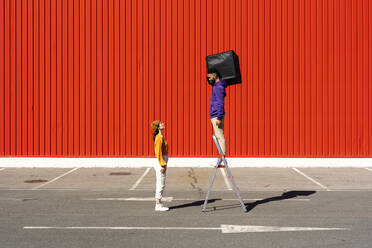 Junger Mann und Frau spielen mit einer Kiste vor einer roten Wand - ERRF02853