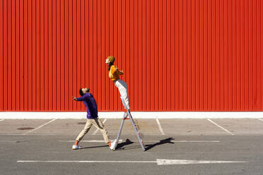 Junger Mann und Frau spielen mit einer Leiter vor einer roten Wand - ERRF02856