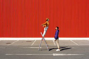 Junger Mann und Frau spielen mit einer Leiter vor einer roten Wand - ERRF02858