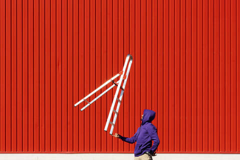 Young man balancing a ladder in front of a red wall stock photo