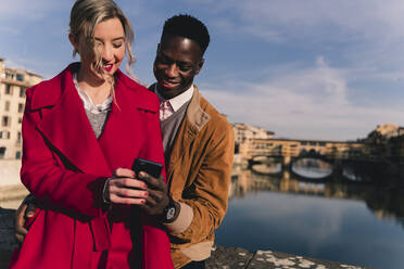 Lächelndes junges Paar teilt sich ein Handy in der Stadt Florenz, Italien - FMOF00876