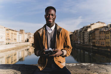 Porträt eines lächelnden jungen Mannes mit einer Kamera auf einer Brücke über dem Fluss Arno, Florenz, Italien - FMOF00883