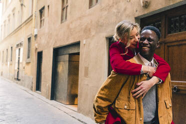 Happy young man carrying girlfriend piggyback in an alley in the city of Florence, Italy - FMOF00891