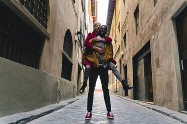 Glücklicher junger Mann, der seine Freundin huckepack in einer Gasse in Florenz, Italien, trägt - FMOF00892