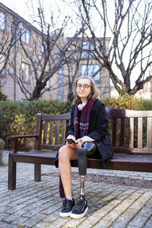 Portrait of confident young woman with leg prosthesis sitting on a bench in the city - FBAF01279