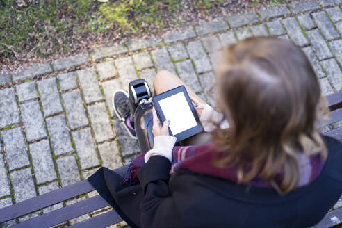 Young woman with leg prosthesis sitting on a bench reading e-book - FBAF01280