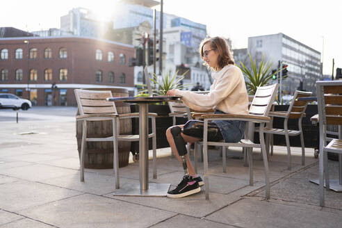 Young woman with leg prosthesis sitting in a sidewalk cafe in the city using laptop - FBAF01282