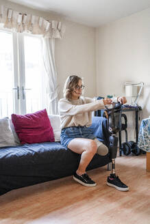 Young woman with leg prosthesis sitting on couch at home - FBAF01289