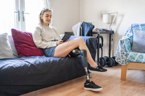 Young woman with leg prosthesis sitting on couch at home reading e-book - FBAF01290