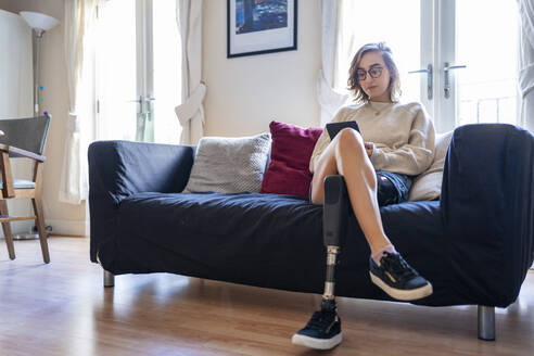 Young woman with leg prosthesis sitting on couch at home reading e-book - FBAF01291