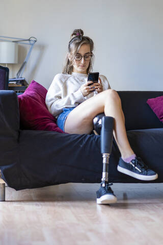 Young woman with leg prosthesis sitting on couch at home using smartphone stock photo