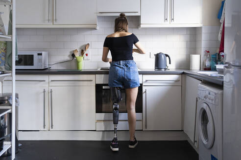 Rear view of young woman with leg prosthesis in kitchen at home - FBAF01295