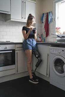 Young woman with leg prosthesis drinking coffee in kitchen at home - FBAF01296