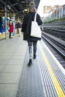 Rear view of young woman with leg prosthesis walking at station platfom - FBAF01298