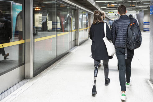 Rear view of young woman with leg prosthesis and man walking at subway station platfom - FBAF01323
