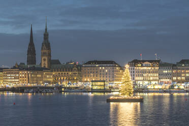 Germany, Hamburg, Illuminated Alstertanne tree at dusk with Jungfernstieg promenade and Saint Nicholas Church in background - KEBF01484