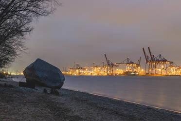 Deutschland, Hamburg, Alter Schwede Findling am Uferstrand in der Abenddämmerung mit Silhouetten von Hafenkränen im Hintergrund - KEBF01516