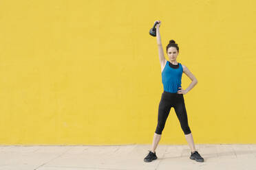 Junge Frau trainiert mit Hantel vor einer gelben Wand - JCZF00023