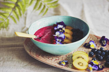 High angle close up of bowl with pink fruit puree with blueberries and slices of yellow kiwis, decorated with pansy petals. - CUF54764