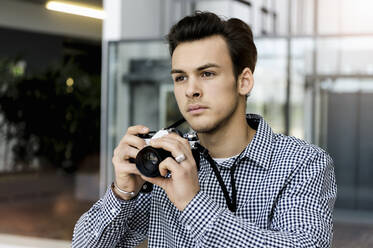 Porträt eines jungen Mannes mit dunkelbraunem Haar, der eine Spiegelreflexkamera hält. - CUF54876