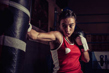 Weibliche Boxerin mit rotem Oberteil trainiert im Fitnessstudio und schlägt auf den Sandsack. - CUF54915