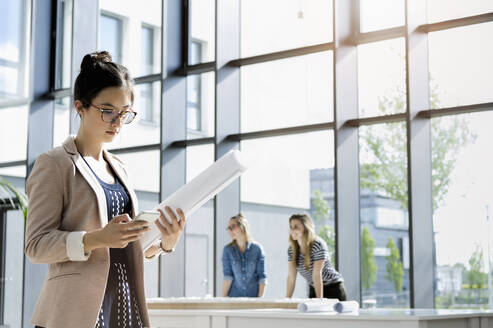 Porträt einer jungen Geschäftsfrau mit dunkelbraunem Haar und Brille, die in einem Büro steht und liest. - ISF23945