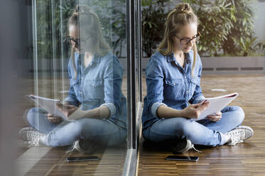 Porträt einer jungen blonden Geschäftsfrau mit Brille, die auf dem Boden sitzt und liest. - ISF23949