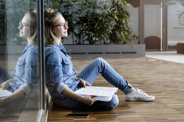 Porträt einer jungen blonden Geschäftsfrau mit Brille, die auf dem Boden sitzt und liest. - ISF23950