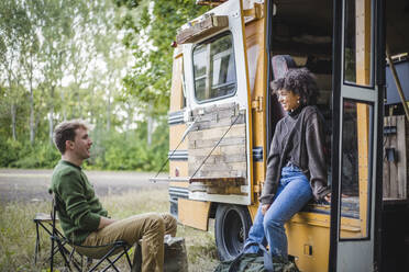 Lächelnde Frau, die sich an die Tür eines Wohnwagens lehnt, während sie sich mit einem männlichen Freund unterhält, der beim Camping im Wald sitzt - MASF16750