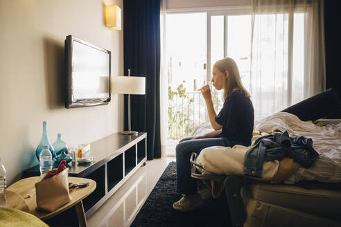 Seitenansicht eines Mädchens, das auf dem Bett eines Hotelzimmers sitzt und fernsieht - MASF16780