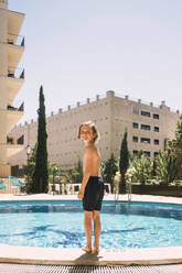 Full length of smiling boy standing by swimming pool against sky - MASF16788