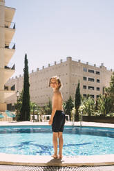 Lächelnder Junge in voller Länge am Swimmingpool gegen den Himmel stehend - MASF16788