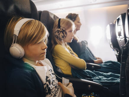 Kaukasischer Junge mit Kopfhörern sitzt mit Schwester und Mutter im Flugzeug - MASF16794