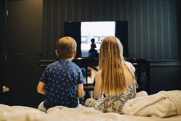 Rear view of siblings watching television while sitting on bed in hotel room - MASF16797