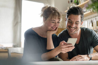 Happy couple sitting at table in kitchen using smartphone - KNSF07650