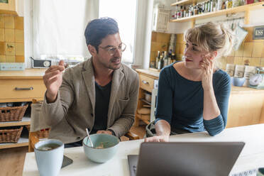 Couple sitting at table in kitchen with laptop talking - KNSF07653