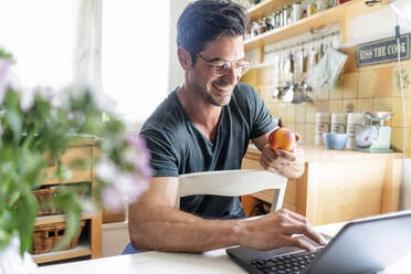 Glücklicher Mann sitzt am Tisch in der Küche und benutzt einen Laptop - KNSF07656
