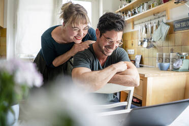 Happy couple in kitchen looking at laptop - KNSF07657