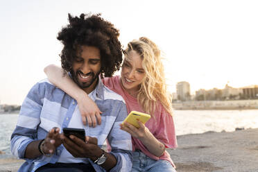 Glückliches junges Paar sitzt bei Sonnenuntergang an der Kaimauer und benutzt sein Smartphone - AFVF05506