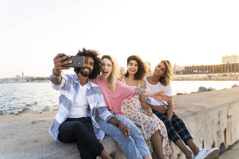 Glückliche Freunde sitzen bei Sonnenuntergang auf der Kaimauer und machen ein Selfie, lizenzfreies Stockfoto